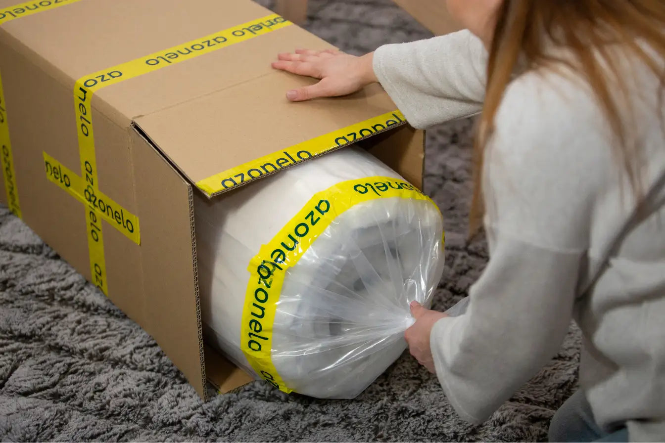Žena vyťahuje zrolovaný matrac Azonelo, zabalený vo fólii so žltou páskou, z kartónovej krabice.