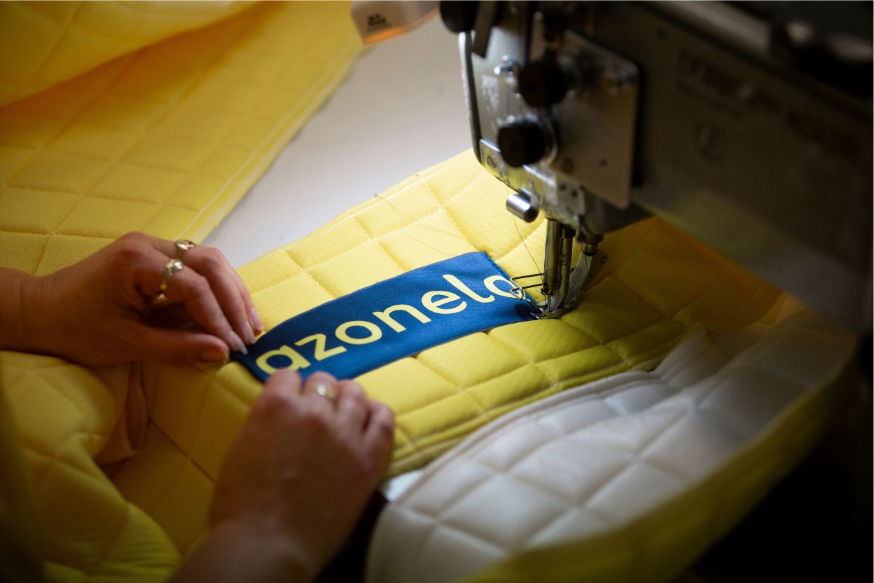 Ručné všívanie modrého loga Azonelo na žltý prešívaný poťah matraca počas výroby na Slovensku. Detailná fotografia zdôrazňuje remeselnú prácu.