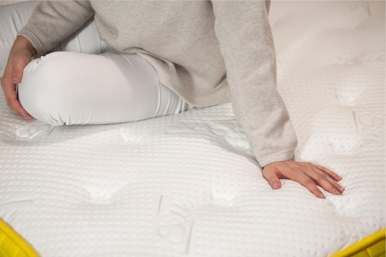 Žena sedí na vysokom matraci Azonelo Cloud s luxusným prešívaným poťahom, testuje jeho pohodlie a nadýchanú štruktúru. Zdôrazňuje komfort a kvalitu matracov Azonelo pre lepší spánok.