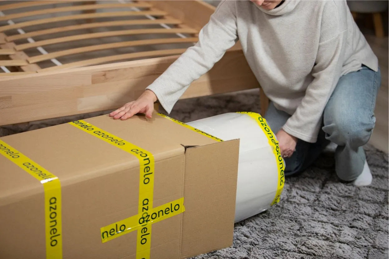 Zákazníčka rozbaľuje vysoký matrac v krabici Azonelo vedľa dreveného roštu, ilustrujúc jednoduché doručenie a inštaláciu